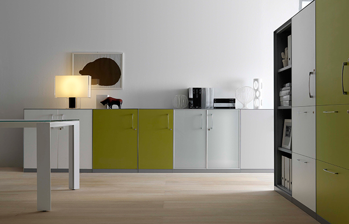Interno di una cucina moderna con armadietti bianchi e verdi, lampada accesa, e scaffale con libri a destra.