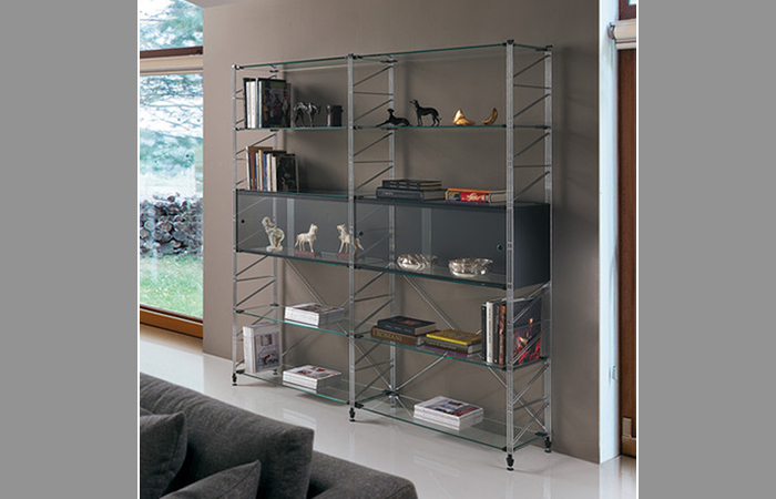 Scaffale moderno in metallo con ripiani in vetro, che ospita libri e oggetti decorativi in un salotto.