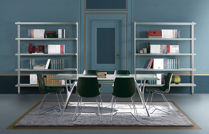 Sala conferenze moderna con pareti blu, tavolo rettangolare bianco e sedie nere, librerie sullo sfondo.