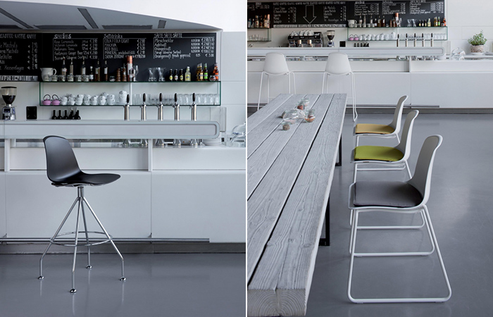 Interno di un bar moderno con arredi bianchi, sedie colorate e un lungo tavolo centrale in legno chiaro.