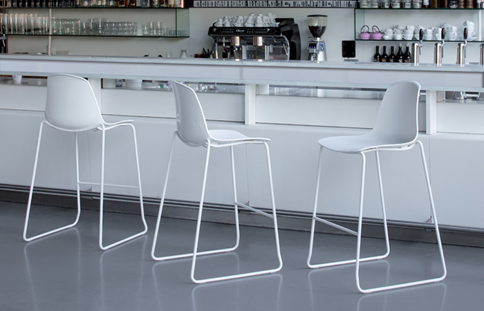 Moderno bar con tre sgabelli bianchi davanti a un bancone bianco, sfondo minimalista e arredamento elegante.